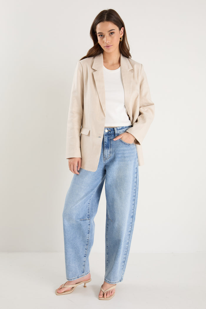 Woman wearing a long-sleeve cream linen blazer with button front and flap pockets, styled with a white top and light blue jeans.