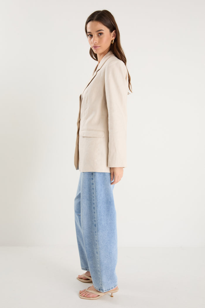 Woman wearing a long-sleeve cream linen blazer with button front and flap pockets, styled with a white top and light blue jeans.