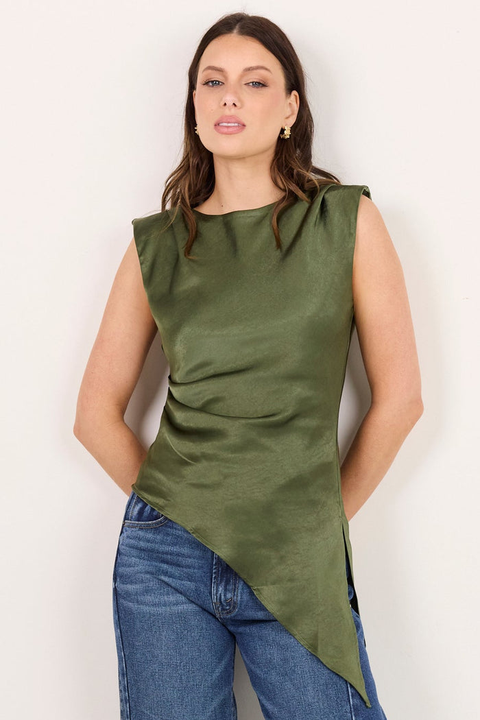 A woman modeling a sleeveless olive green asymmetrical top paired with blue jeans and black sandals, standing against a plain off-white studio background.