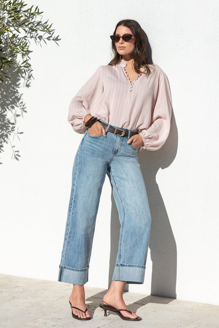 A woman stands against a white wall wearing a light pink long-sleeve blouse, light blue wide-leg jeans, a black belt, and black sandals.