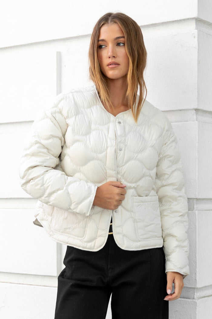 Woman wearing a white puffer jacket and black pants standing against a white wall.
