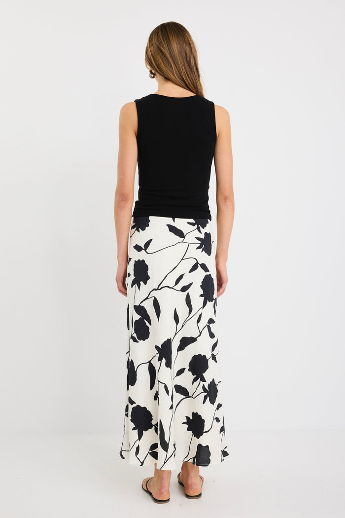 Woman wearing an ivory bias-cut midi skirt with black floral print, elastic waist, and pull-on fit, styled with black sandals and a tucked-in black top.