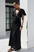 A woman wearing a black maxi dress with white contrast piping stands outdoors in front of a modern white building with columns.