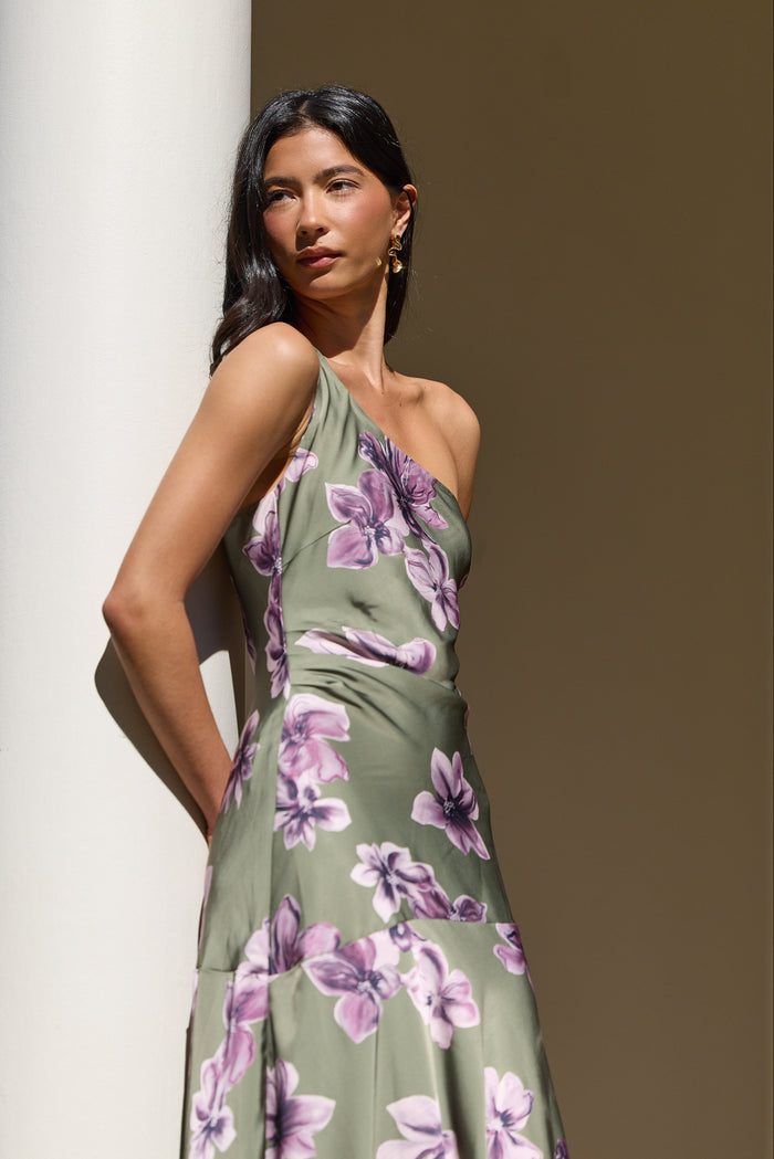 A woman wearing a one-shoulder olive green dress with a purple floral print, posing next to a white column in warm, natural light.