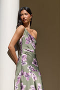 A woman wearing a one-shoulder olive green dress with a purple floral print, posing next to a white column in warm, natural light.