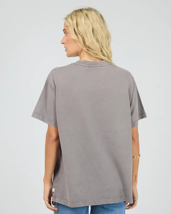 A woman with blonde hair is shown from the back wearing an oversized, washed gray t-shirt and blue jeans against a light gray studio background.