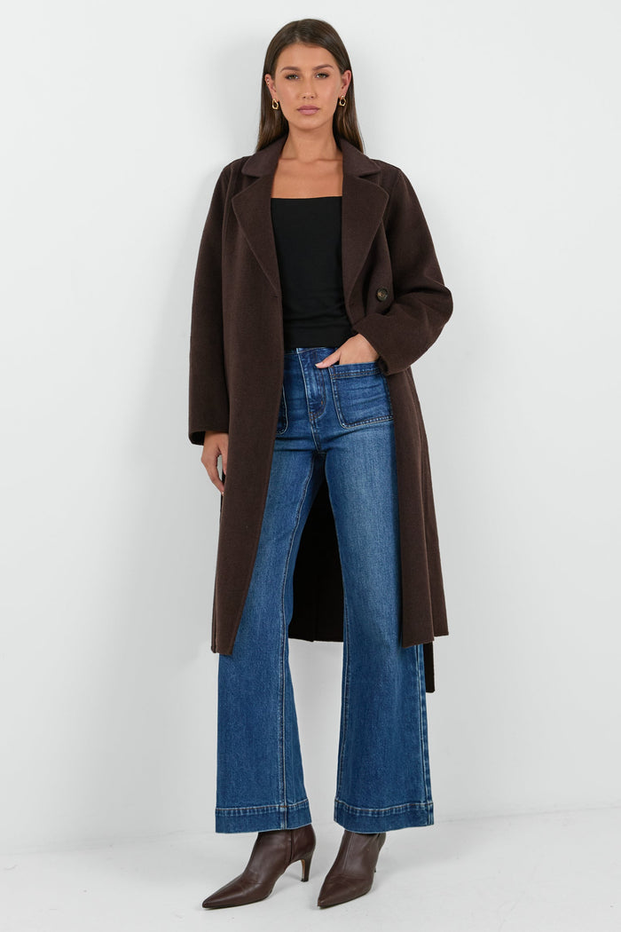 Woman wearing a brown coat, black top, and blue jeans on a white background