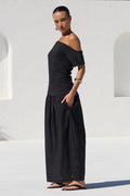 A woman wearing a black off-the-shoulder maxi dress stands on a white tiled poolside patio with a modern white stucco building in the background.