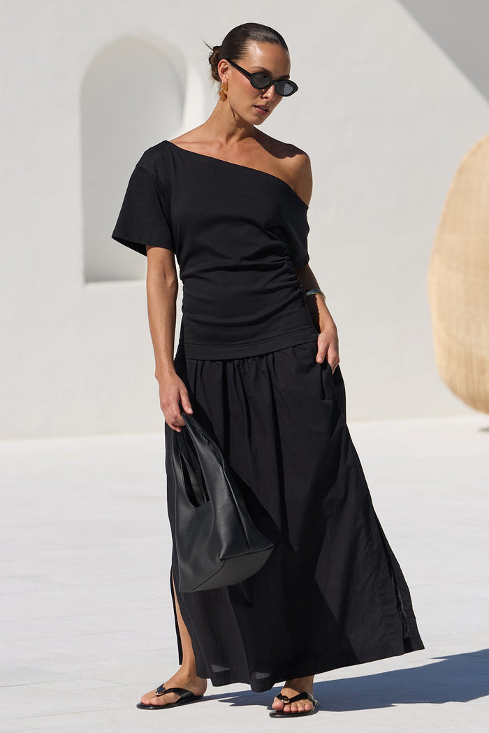 A woman wearing a black off-the-shoulder dress stands on a white patio poolside, holding a black handbag. The background features modern white architecture, a hanging woven chair, and a blue tiled swimming pool.