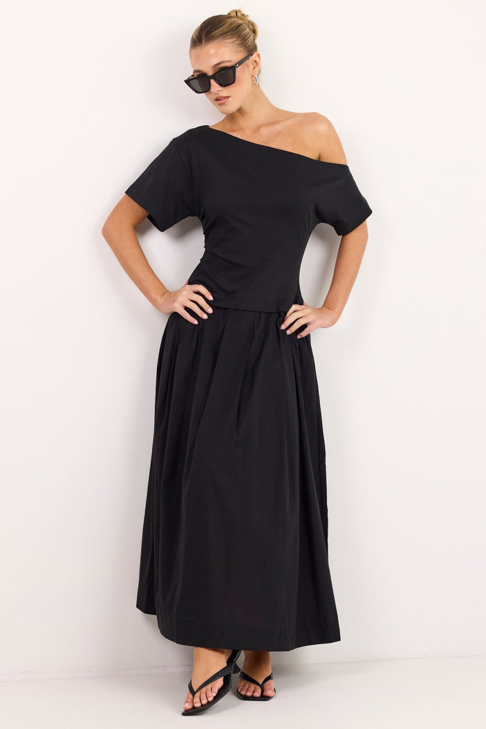 A woman modeling a black off-the-shoulder maxi dress with short sleeves and a flared skirt, paired with black strappy heels and sunglasses, standing against a plain white studio background.