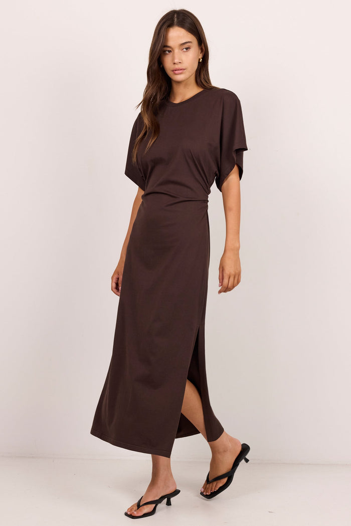 A woman modeling a dark brown short-sleeve maxi dress with a high side slit, paired with black heeled sandals, against a plain off-white studio background.