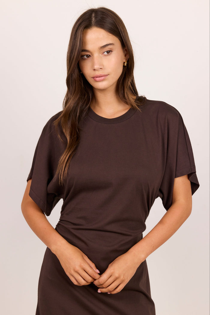 A woman modeling a dark brown short-sleeve t-shirt dress with draped sleeves against a light beige studio background.