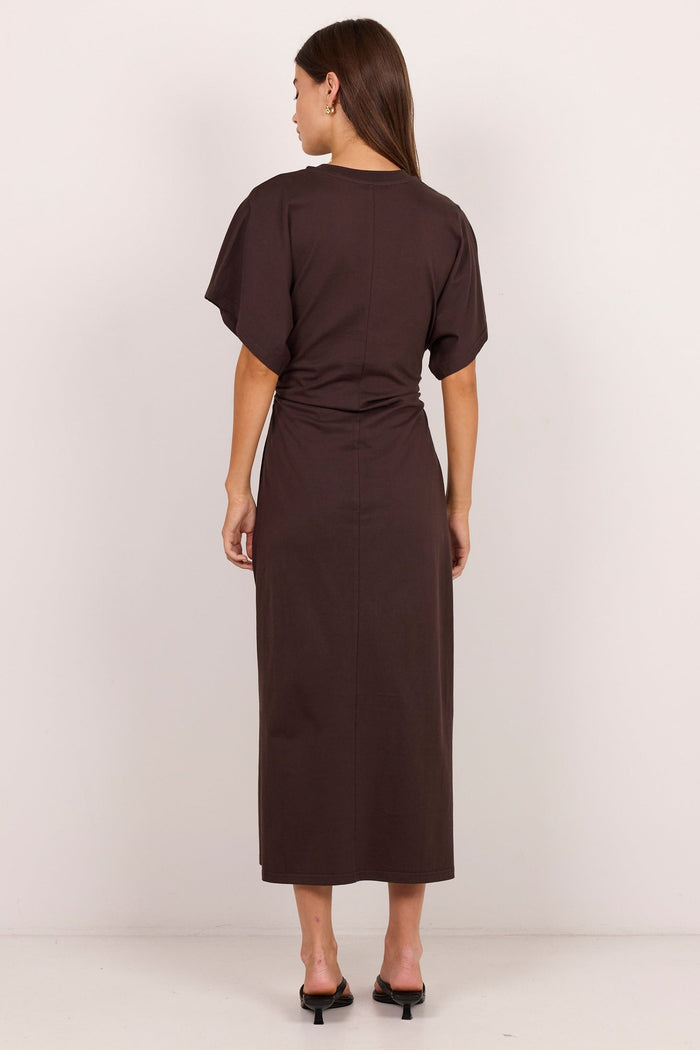 A woman viewed from the back wearing a dark brown short-sleeve midi dress with a gathered waist, paired with black mule heels, against a plain off-white studio background.