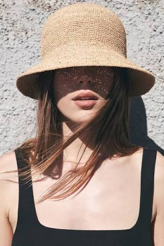 A woman wearing a natural straw bucket hat and a black square-neck top, standing against a textured gray wall with dappled sunlight and shadows.