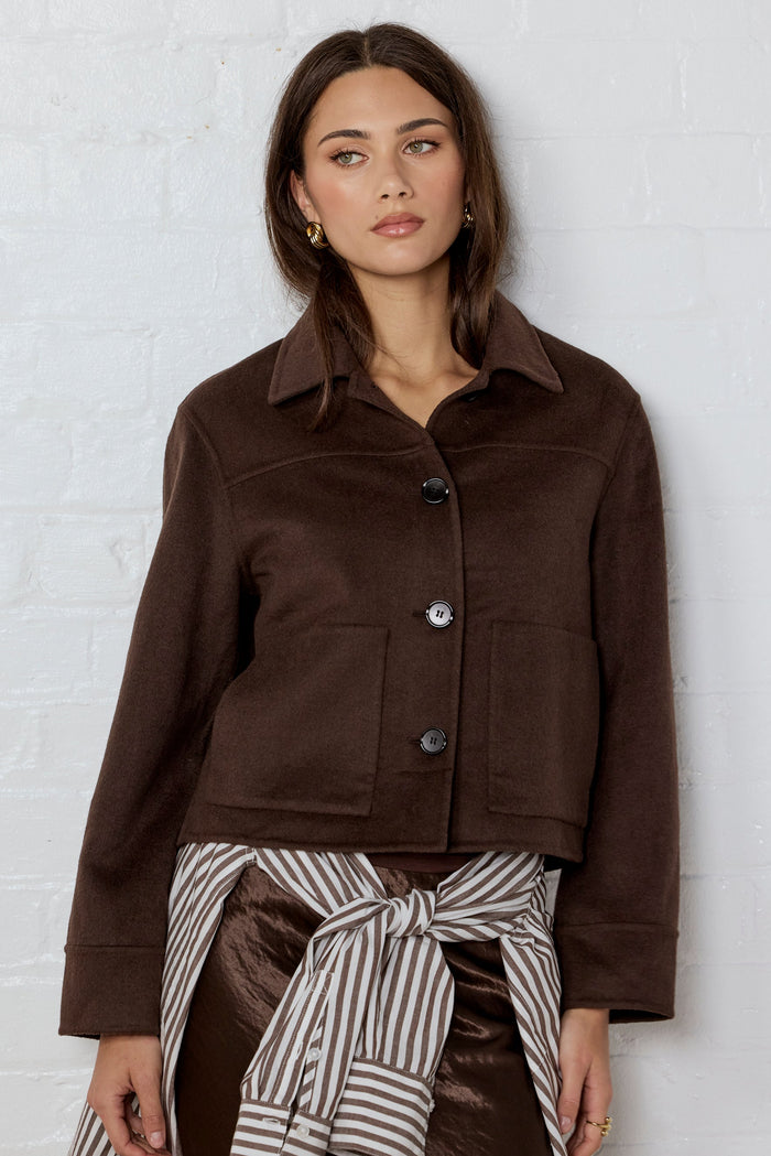 Woman wearing a brown jacket and striped skirt against a white brick wall.