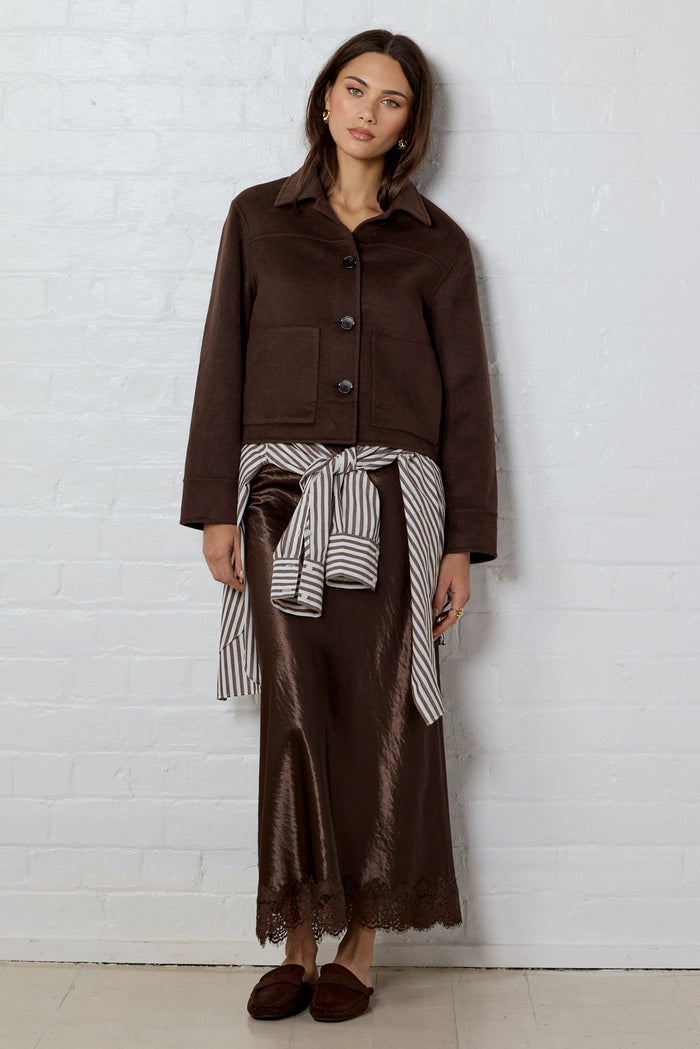 Woman wearing a brown wool jacket with a satin brown skirt leaning on a white brick wall background