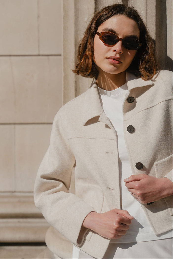 Woman wearing a beige coat and sunglasses against a stone wall.