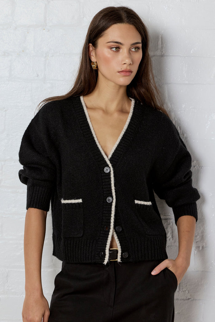 Woman wearing a black cardigan with white trim against a white brick wall.