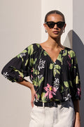 A woman modeling a black floral blouse with green and pink botanical print, paired with white shorts, standing against a light wall with strong sunlight and shadows.