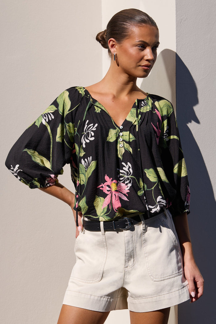 A woman models a black blouse with a green and pink tropical floral print, paired with light beige shorts, standing against a minimalist beige wall with a strong diagonal shadow.