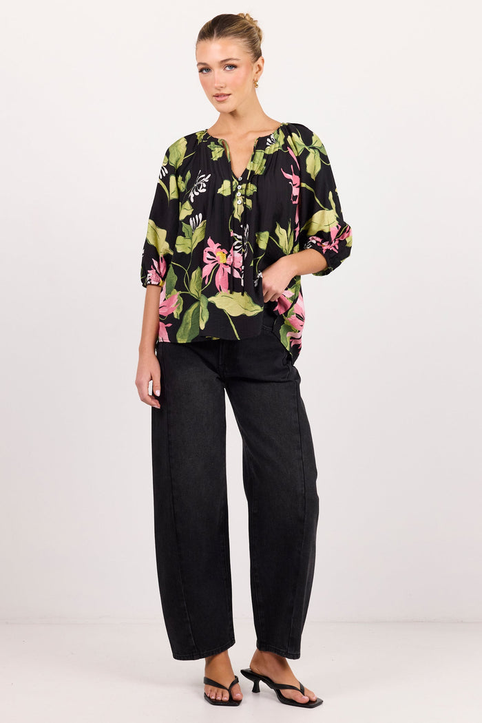 A woman modeling a black three-quarter sleeve blouse with a vibrant green, pink, and white tropical floral print, paired with black wide-leg trousers and black heeled sandals, against a plain white studio background.
