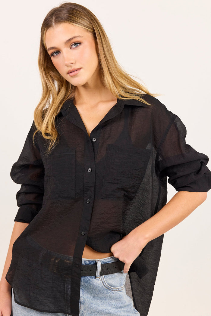 A woman wearing a black sheer button-up shirt with textured side panels, paired with light-wash jeans and a black belt, against a plain off-white background.