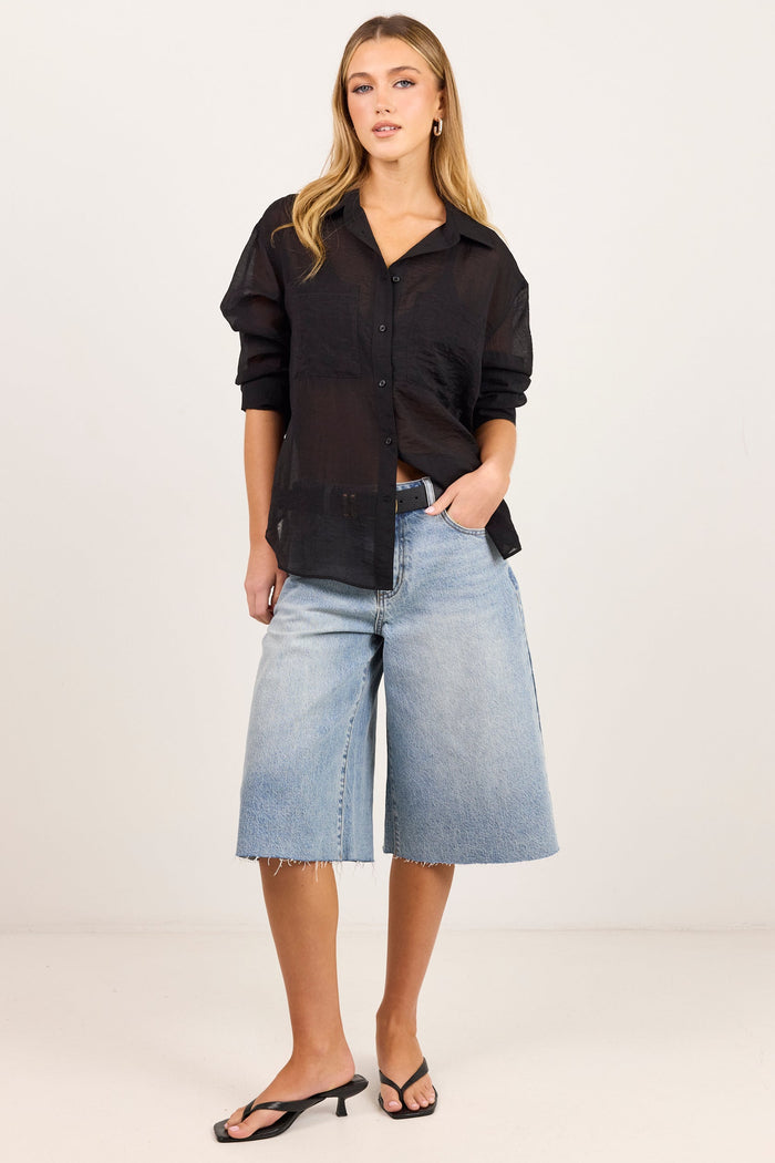 A woman modeling a black sheer long-sleeve button-up shirt paired with light-wash denim culotte shorts and black heeled sandals, standing against a plain off-white studio background.