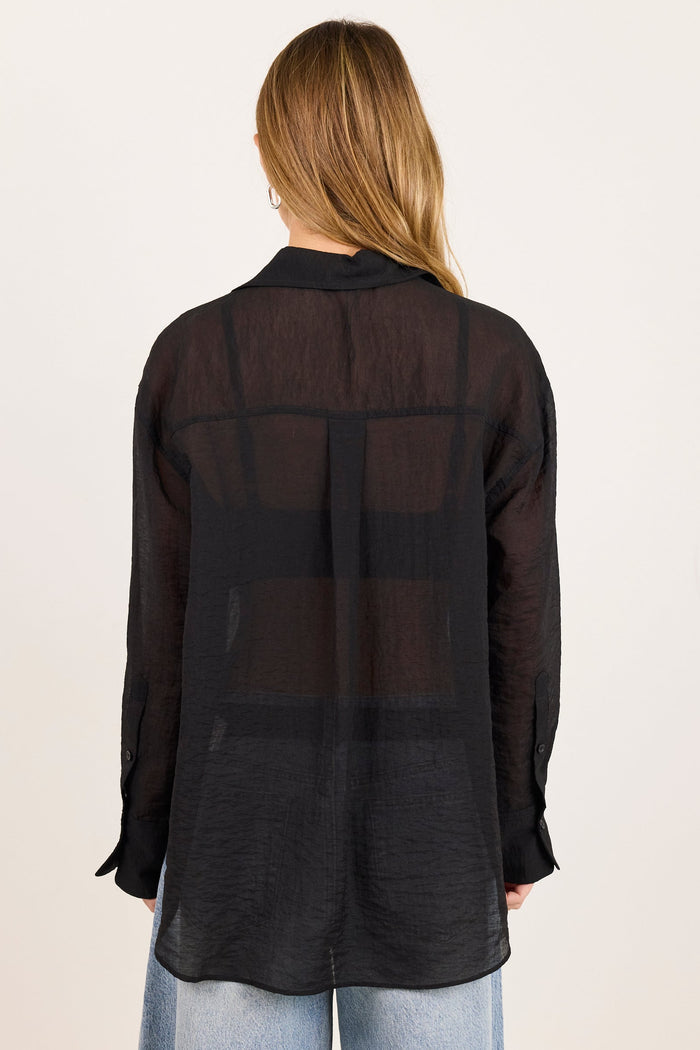 A woman viewed from behind wearing a sheer black long-sleeve button-up shirt over light blue jeans, set against a plain off-white studio background.