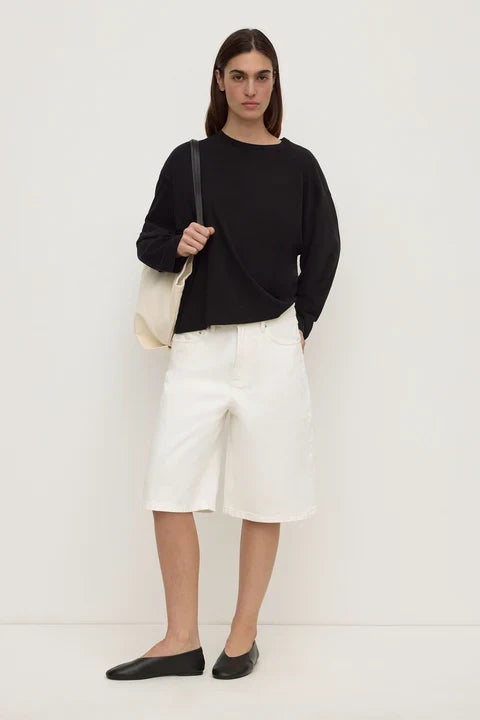 A woman stands against a plain off-white studio wall wearing a black long-sleeve top, white knee-length shorts, and black flats, while carrying a cream shoulder bag.
