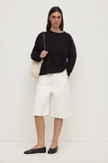 A woman stands against a plain off-white studio wall wearing a black long-sleeve top, white knee-length shorts, and black flats, while carrying a cream shoulder bag.