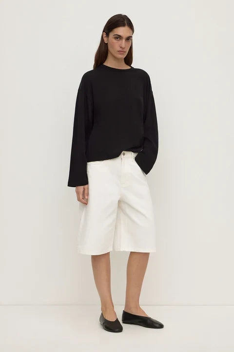 A model stands against a plain white studio background wearing a black long-sleeve top, white cropped shorts, and black flats.