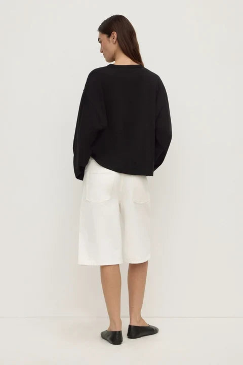 A woman shown from the back wearing a black long-sleeve cropped sweater, white wide-leg shorts, and black flats, standing against a plain white studio background.