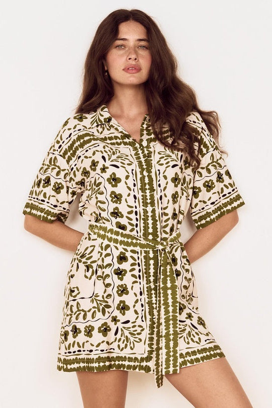 A woman modeling a short-sleeve belted shirt dress with an olive green and cream block print pattern against a plain white background.