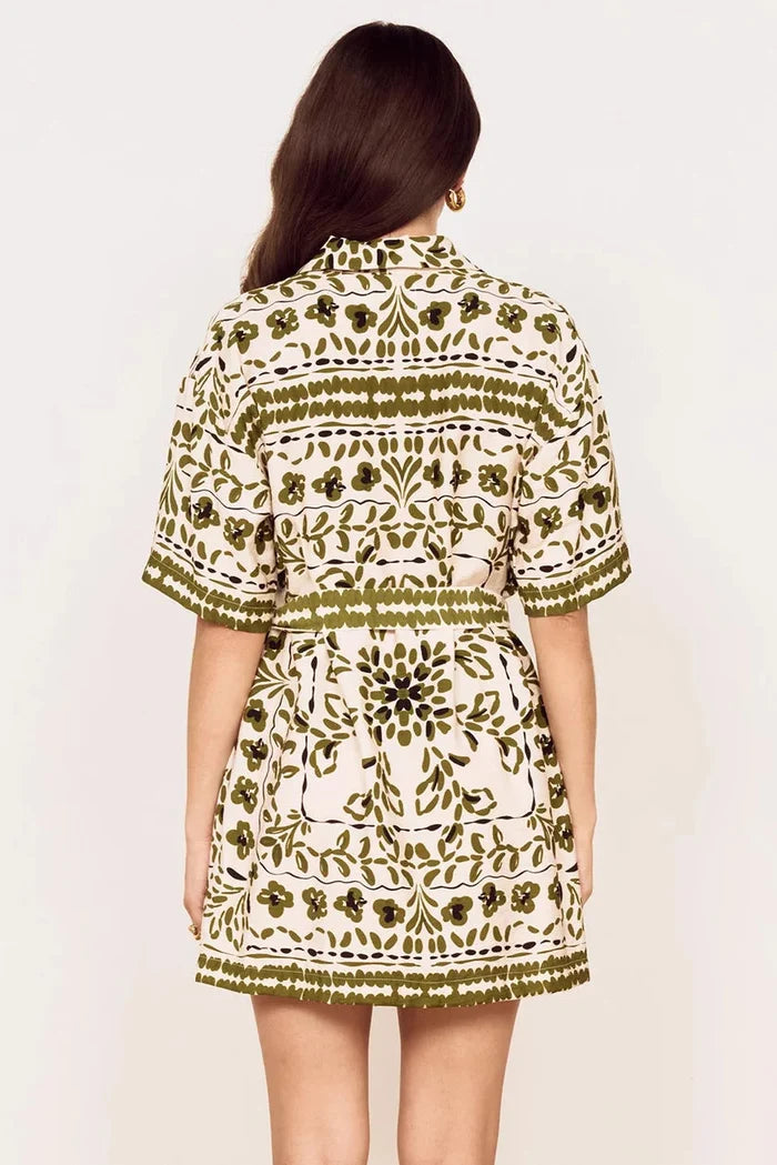 A woman viewed from the back wearing a short-sleeve collared dress with an olive green and cream floral block-print pattern, set against a plain off-white background.