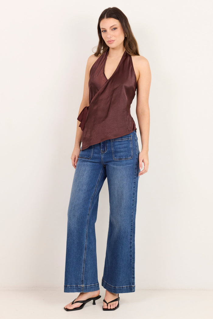 A woman modeling a dark brown halter wrap top and blue wide-leg jeans against a plain white studio background.