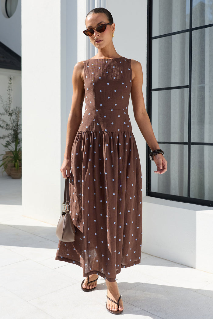 A woman models a sleeveless brown maxi dress with a blue polka dot pattern, walking outdoors on a white tiled patio next to a white building with a large black-framed window.
