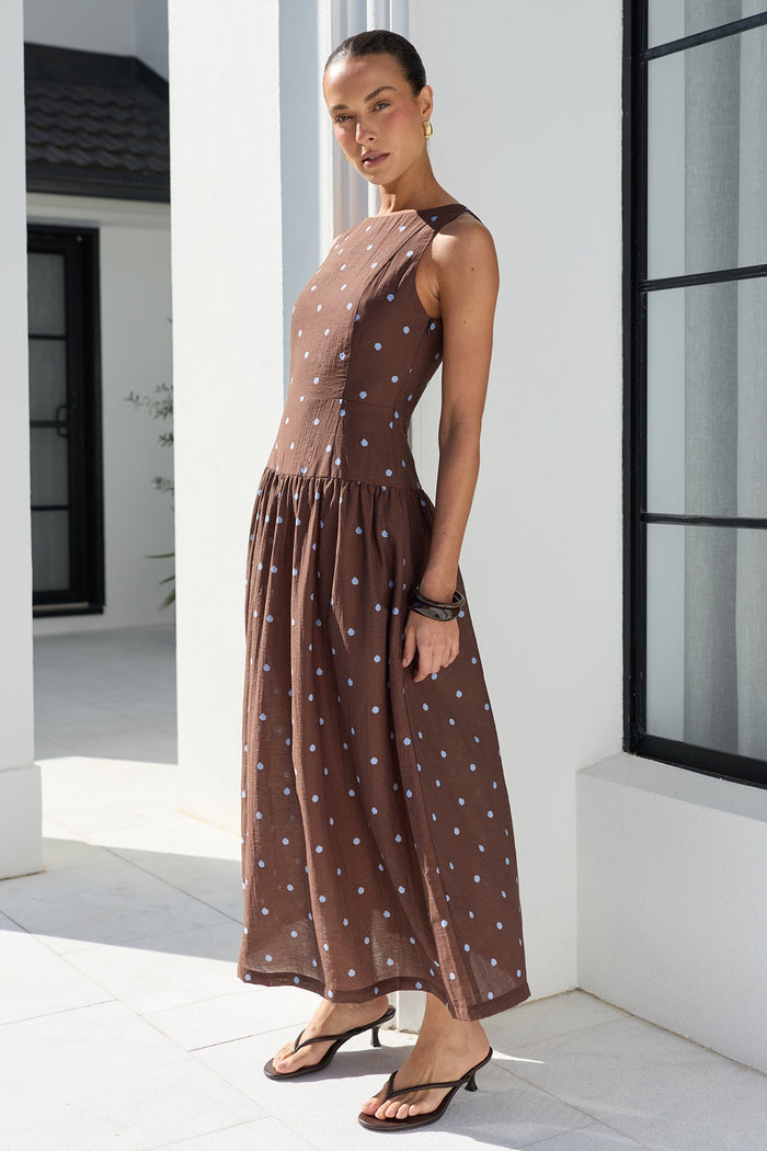 A woman modeling a sleeveless brown maxi dress with a blue polka dot pattern, standing in a bright, modern white architectural setting with a large black-framed window.