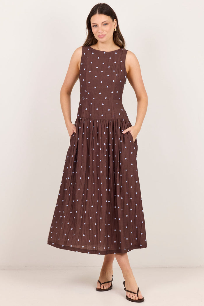 A woman modeling a sleeveless brown midi dress with a white polka dot pattern, standing against a plain light beige studio background.
