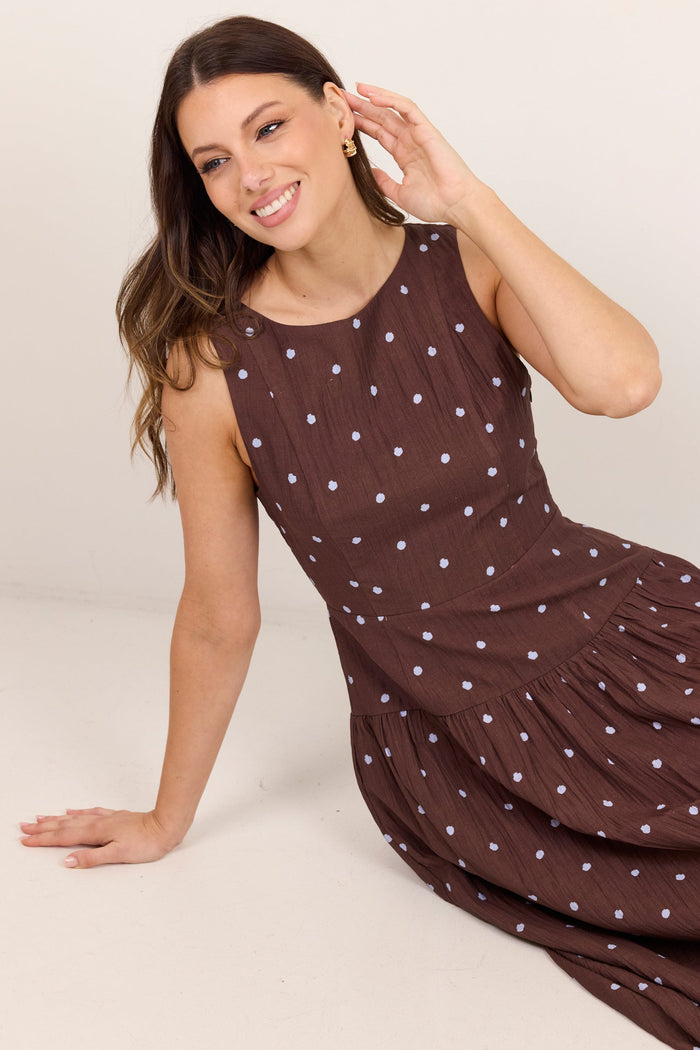 A woman wearing a sleeveless brown dress with a white polka-dot pattern, posing on a light beige studio background.