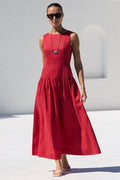A woman wearing a sleeveless red midi dress, sunglasses, and a long necklace walks by a white poolside next to a white building with arched windows.