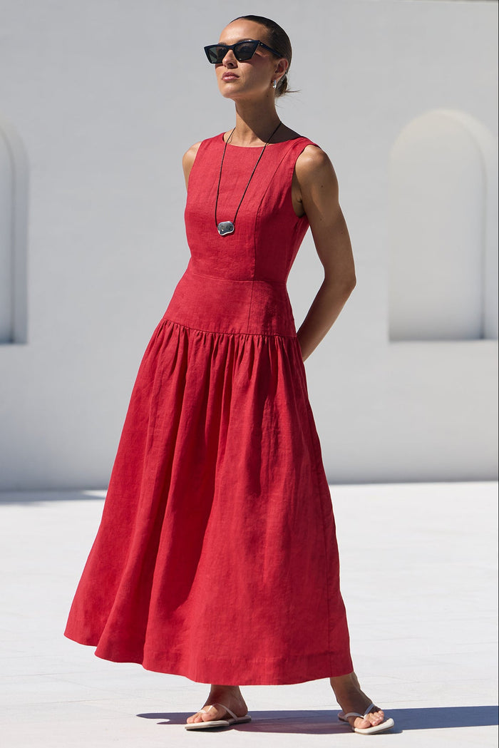 A woman wearing a sleeveless red maxi dress and sunglasses stands on a white tiled poolside deck with a modern white building and trees in the background.