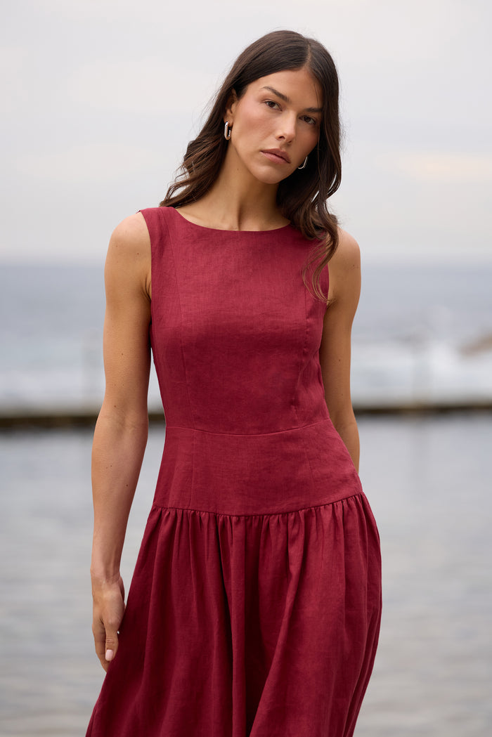 A woman wearing a sleeveless, deep burgundy dress with a textured linen-like fabric, standing outdoors near a body of water with a soft-focus background.