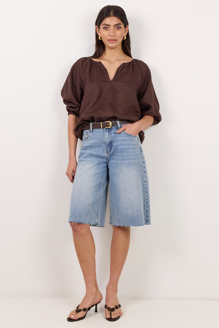 A woman modeling a dark brown long-sleeve blouse and light-wash denim cropped shorts with a black belt, standing against a plain white studio background.