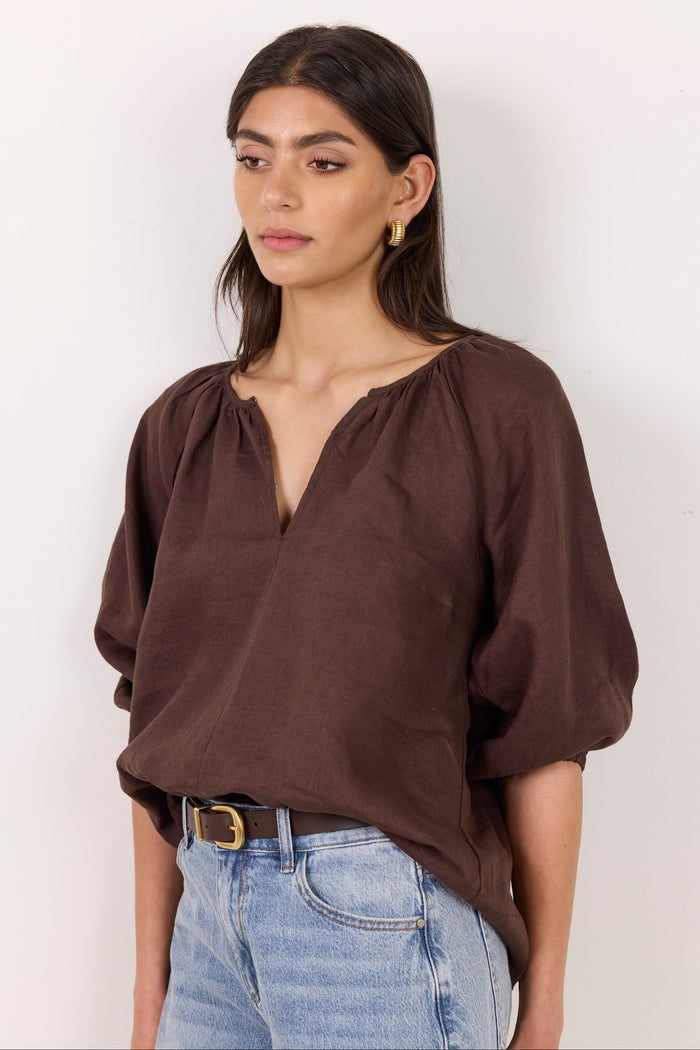 A woman wearing a dark brown, loose-fitting linen blouse with a split neckline and voluminous elbow-length sleeves, paired with light-wash blue jeans and a brown belt, set against a plain off-white background.