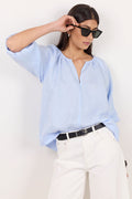 A woman wearing a light blue linen blouse, white trousers, and black sunglasses, posing against a plain white wall.