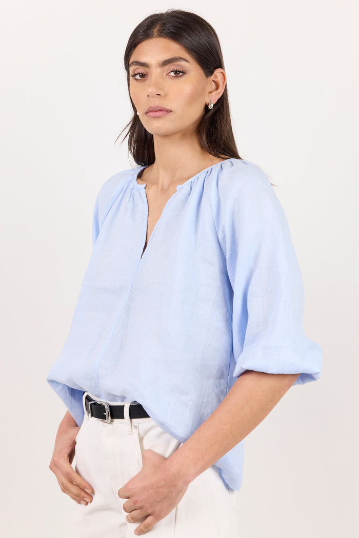 A woman modeling a light blue linen blouse with a split neckline and elbow-length sleeves, paired with white trousers and a black belt, against a plain white studio background.