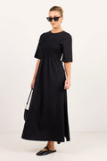 A woman modeling a black midi dress with short sleeves, a round neckline, and a smocked waist, styled with black sunglasses, black flats, and a small black handbag against a plain white studio background.