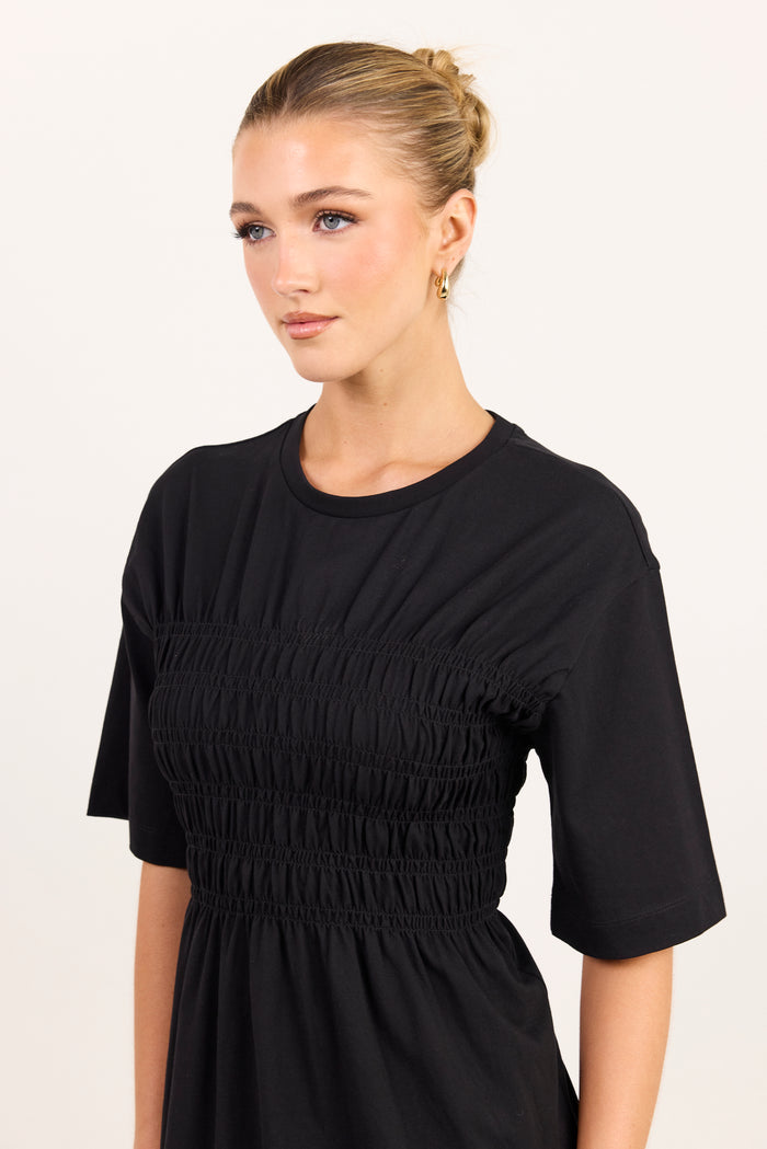 A woman wearing a black short-sleeve dress with a smocked bodice, photographed against a plain light background.