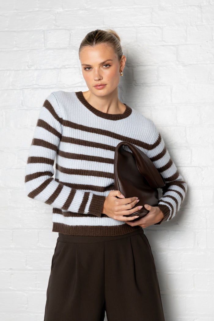 Woman wearing a brown and white striped sweater holding a clutch against a white brick wall.