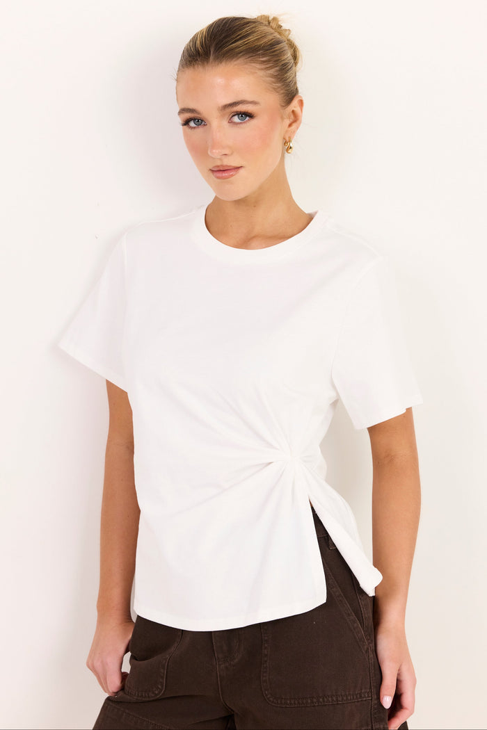 A woman modeling a white short-sleeve t-shirt with a twisted knot detail at the side hem, paired with dark brown pants, against a plain white background.